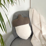 Gray armchair with a white acorn cushion and brown cover, draped with a beige blanket, next to a plant.