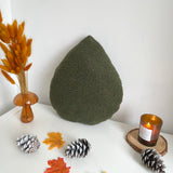 Green textured teardrop-shaped pillow on a white surface with decorative elements like a candle, leaves, and pinecones.