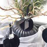 Black ornament with a silver bow on a branch with white fabric and greenery in the background