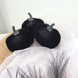 Three black velvet pumpkins on a white surface with a straw hat and decorative branches.