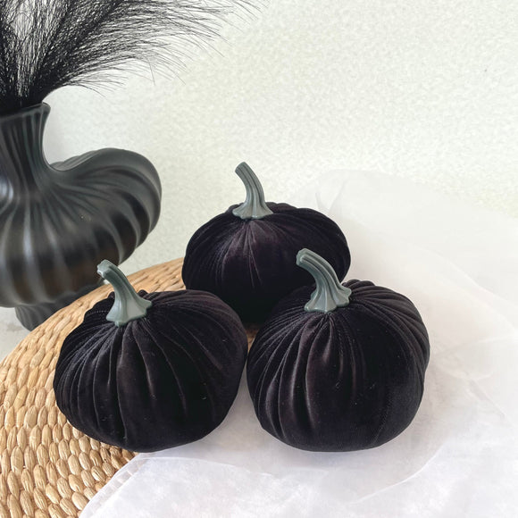 Black velvet pumpkins on a white surface with a woven mat and decorative vase.