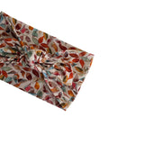 A knotted fabric headband with a multicolor autumn leaf pattern on a cream background, shown diagonally on a plain white background.