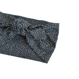 A close-up of a knotted fabric headband with a black leopard print pattern on a muted blue-gray background, shown against a plain white background.