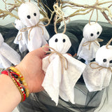 Hand holding a small white ghost decoration with black eyes and a bow, surrounded by more ghost decorations.