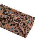 A knotted fabric headband with a leopard print pattern in brown, black, and multicolor accents, shown on a plain white background.
