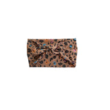A leopard print fabric headband with a large twisted knot at the front, featuring a tan base with black spots and small multicolor accents, shown on a plain white background.