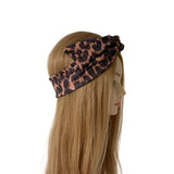 A leopard print fabric headband with a large bow, displayed on a mannequin head with long blonde hair against a plain white background.