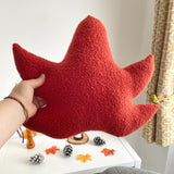 Red leaf-shaped pillow held by a hand on a white surface with decorative items.