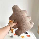 Brown textured leaf-shaped pillow held by a hand on a white surface with decorative items.