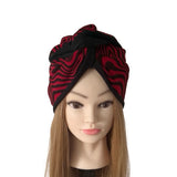 A red and black patterned fabric headwrap turban hat displayed on a mannequin head with long light brown hair against a plain white background.