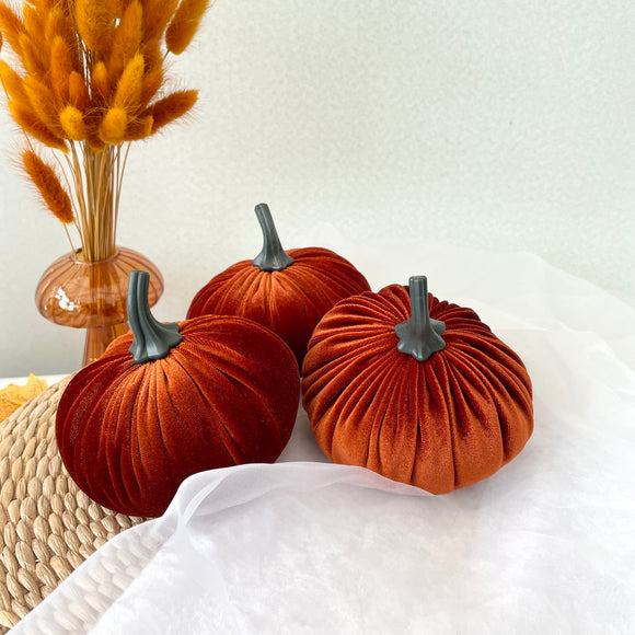 Three orange velvet pumpkins with gray stems on a white surface, with a vase of dried grasses in the background.