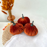 Three orange velvet pumpkins on a white surface with a woven mat and dried grasses in the background.
