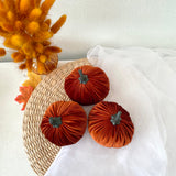 Three rust-colored velvet pumpkins with green stems on a woven mat with a white cloth and dried flowers in the background.