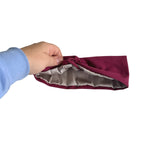 A hand wearing a light blue long-sleeve shirt holds open a burgundy drawstring pouch, showing a shiny silver-gray satin lining, against a plain white background.