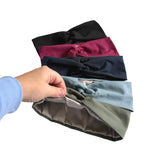 A hand wearing a light blue long-sleeve shirt holds a stack of four twisted fabric headbands in black, burgundy, navy, and sage green against a plain white background.