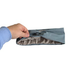 A hand wearing a light blue long-sleeve shirt holds a folded dusty blue fabric pouch or sleeve with a gray interior lining against a plain white background.