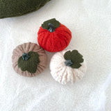 Three boucle fabric mini pumpkins in rust, taupe, and cream white on a textured white surface.