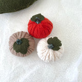 Three boucle fabric mini pumpkins in rust, taupe, and cream white on a textured white surface.