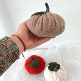 Hand holding a plush brown pumpkin with two other pumpkins, one red and one white, on a white background.