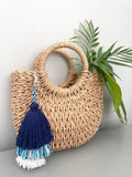 A handmade blue and white tiered tassel keychain placed on a woven tote bag, with a green tassel adding a decorative touch to the beige background.