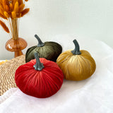 Four velvet pumpkins in red, green, and gold on a white surface with a vase of dried grasses in the background.