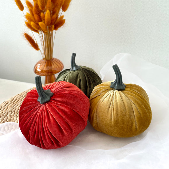 Three velvet pumpkins in rust, green, and gold on a white surface with a vase of dried grasses in the background.