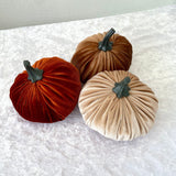 Three velvet pumpkins in orange, brown, and beige on a textured white surface.
