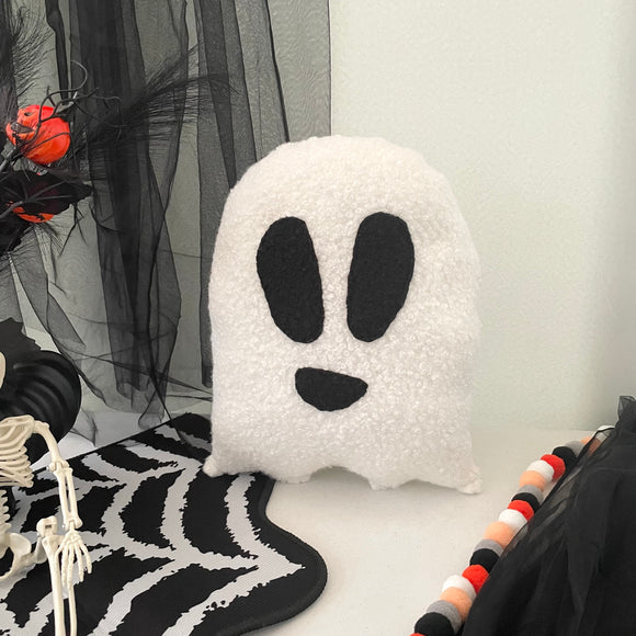 Ghost-shaped pillow on a Halloween-themed background with spider webs and skeleton.