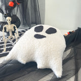White ghost-shaped pillow with black spots on a Halloween-themed background with skeleton and decorations.