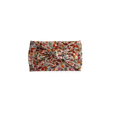 A fabric headband with a large twisted knot, featuring an all-over autumn leaf pattern in shades of orange, red, brown, and green on a light beige background.