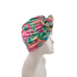 A multicolor floral turban hat with pink, green, and peach tones, displayed on a white mannequin head in profile against a white background.