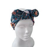 A dark teal blue floral turban headband with a large bow, displayed on a white mannequin head against a solid white background.