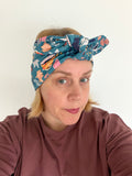 A woman wearing a teal blue patterned headband with a twisted knot, paired with a dusty rose long-sleeve shirt, photographed against a plain off-white background.