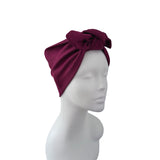 A deep burgundy ribbed fabric headwrap turban with a large bow on top, displayed on a white mannequin head against a plain white background.