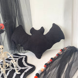 Black bat-shaped pillow on a white surface with Halloween decorations.