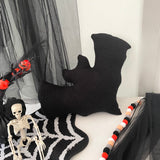 Black bat-shaped pillow on a white surface with Halloween decorations including a skeleton and garlands.