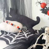 Black cat plush toy on a white pillow with Halloween decorations including a skeleton and pumpkins.
