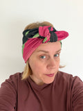 A woman wearing a knotted fabric headband with pink, green, and black stripes, paired with a brown long-sleeve top against a plain white background.