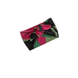 A pleated fabric headband with a large bow, featuring a geometric pattern of black, magenta pink, and green, shown on a plain white background.
