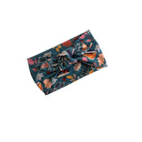 A fabric headband with a large twisted knot, featuring a detailed botanical and butterfly pattern in teal, pink, orange, and green on a solid white background.
