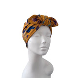 A mustard yellow fabric headwrap turban with a purple and blue floral pattern, displayed on a white mannequin head against a white background.