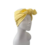 A yellow ribbed fabric headwrap turban hat with a large twisted knot on top, displayed on a white mannequin head against a plain white background.