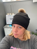 Person wearing a black beanie with a fluffy pom-pom, standing indoors.