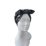 A black shiny fabric headwrap or turban with a large bow on top, displayed on a white mannequin head against a plain white background.