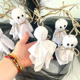 Hand holding small white ghost decorations with black eyes and brown ribbons against a wreath background.