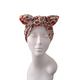 A multicolor floral print headwrap styled on a white mannequin head, featuring a large bow at the top and a subtle leaf pattern in orange, red, blue, and green on a cream background.