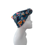 A teal blue fabric headwrap or turban hat with a colorful floral and botanical print, displayed on a white mannequin head in profile against a plain white background.
