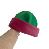 A two-tone fleece beanie hat held by a person's hand. The top half is green, and the folded cuff is a deep magenta pink.