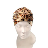 A ruffled fabric headwrap or turban hat with an abstract marbled pattern in beige, brown, black, and orange, displayed on a white mannequin head against a white background.