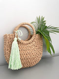 A handmade light green tassel keychain with braided string, displayed against a neutral background with a wicker bag and a green plant.